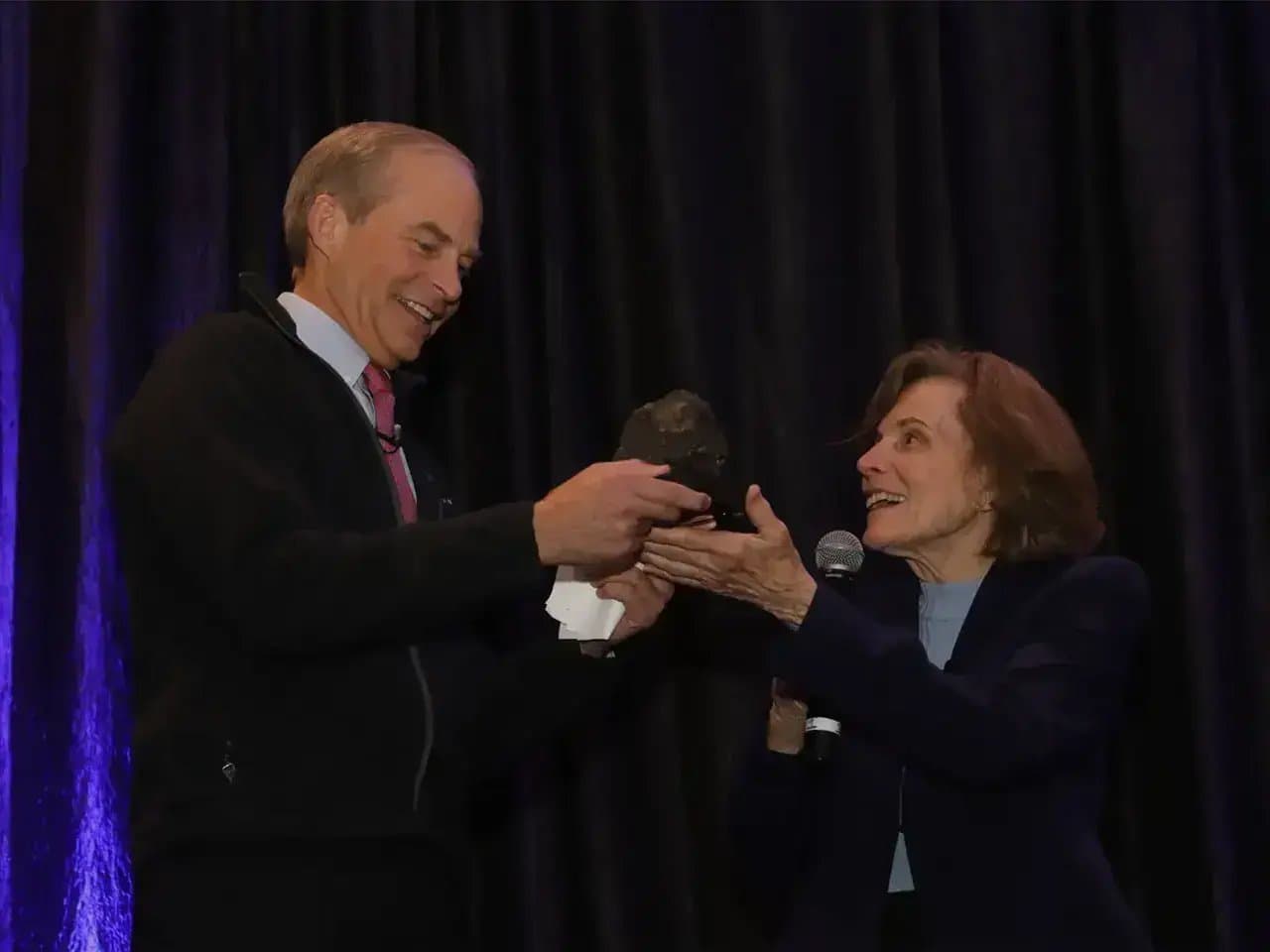 A man hands an object to a woman holding a microphone, both smiling, in front of dark curtains at an indoor event.