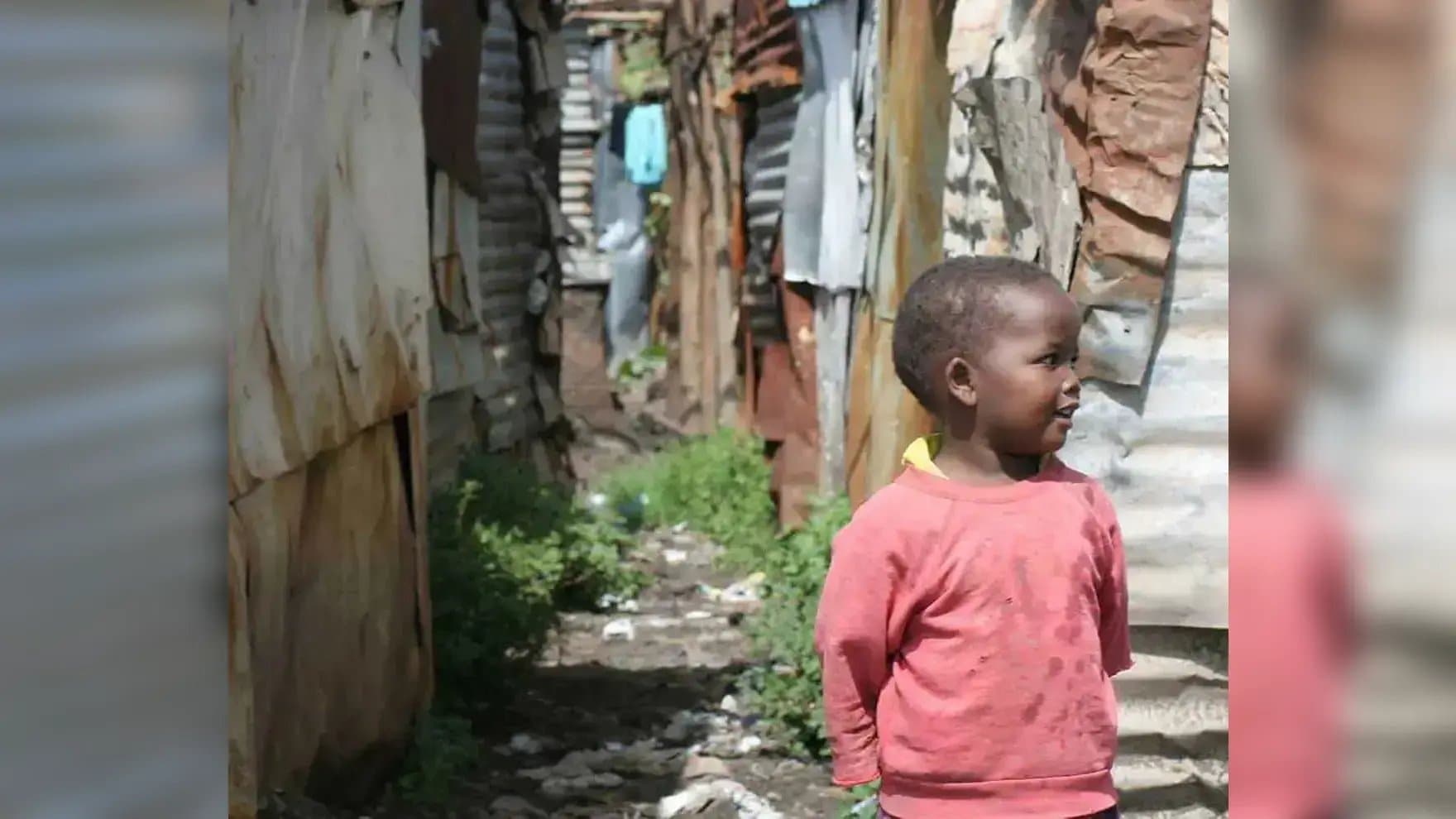 A young child stands smiling, wearing a pink sweater, in an alley between makeshift metal and wood structures. The pathway is dirt-covered with patches of green vegetation.