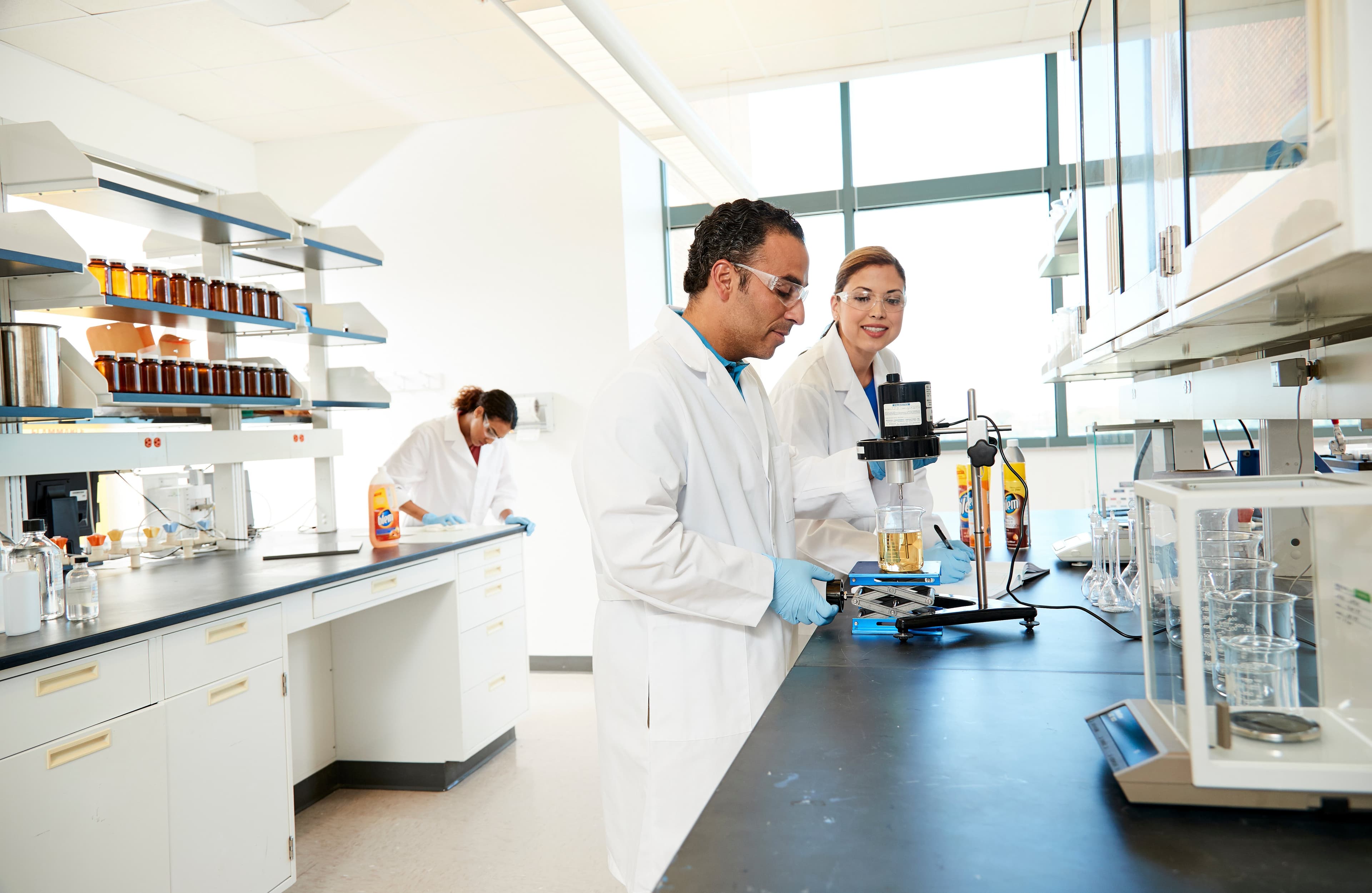 Scientists in lab coats conduct experiments with liquids using equipment in a bright, organized laboratory. Shelves with jars are visible, indicating a setting of research and analysis.