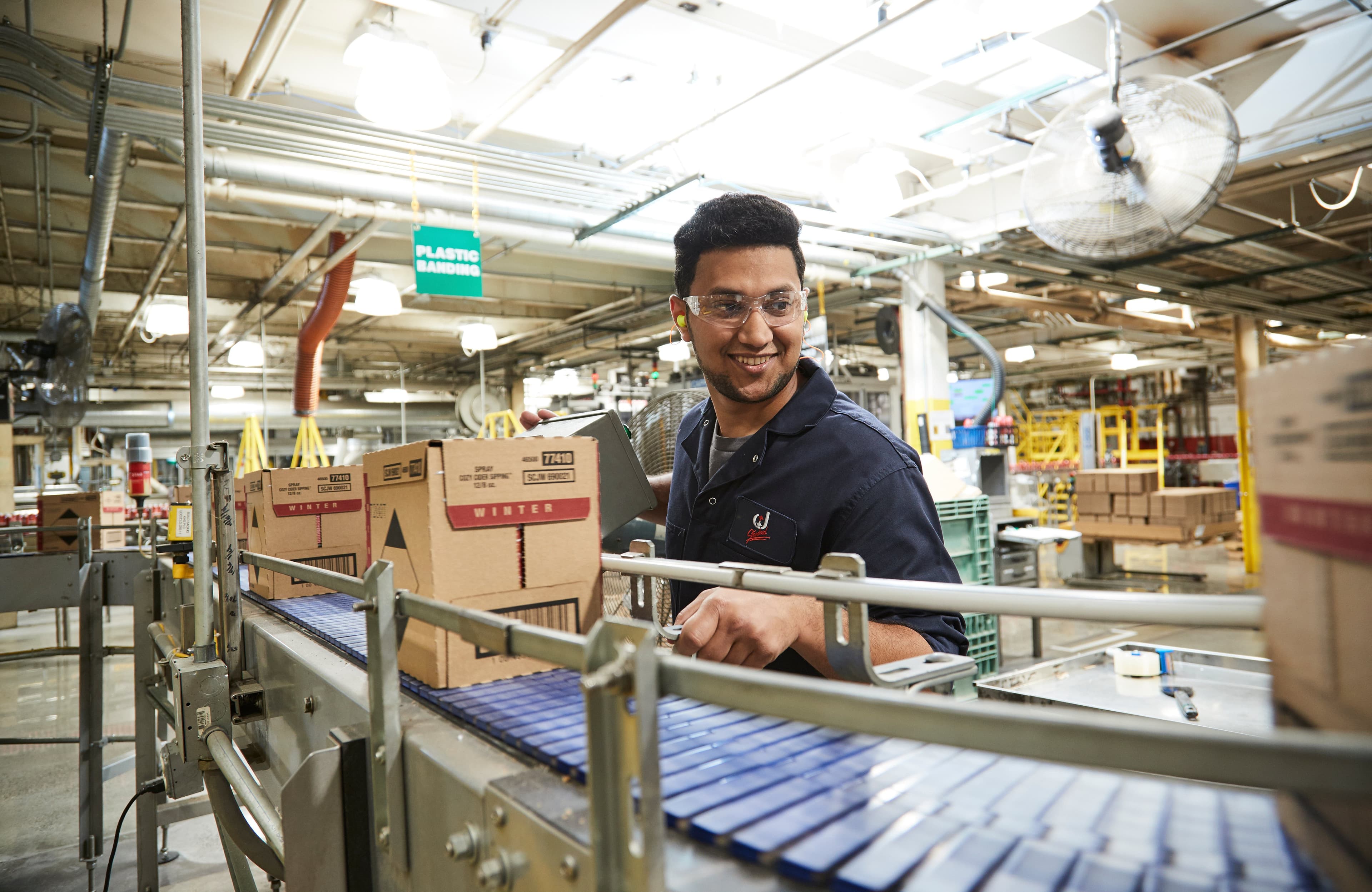 A worker moves a box along a conveyor belt in a factory setting. Overhead, a sign reads "PLASTIC BANDING." The environment is industrial, with machinery and bright lighting.