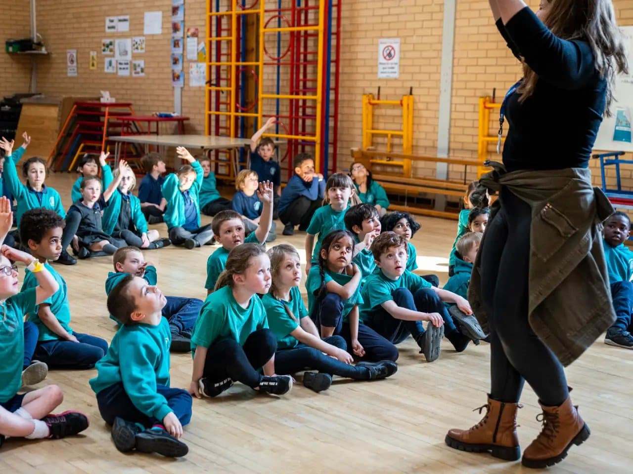 Children sit on a gym floor, some raising hands, attentively watching a standing adult. The setting includes gym equipment and stacked chairs against a brick wall with posters.
