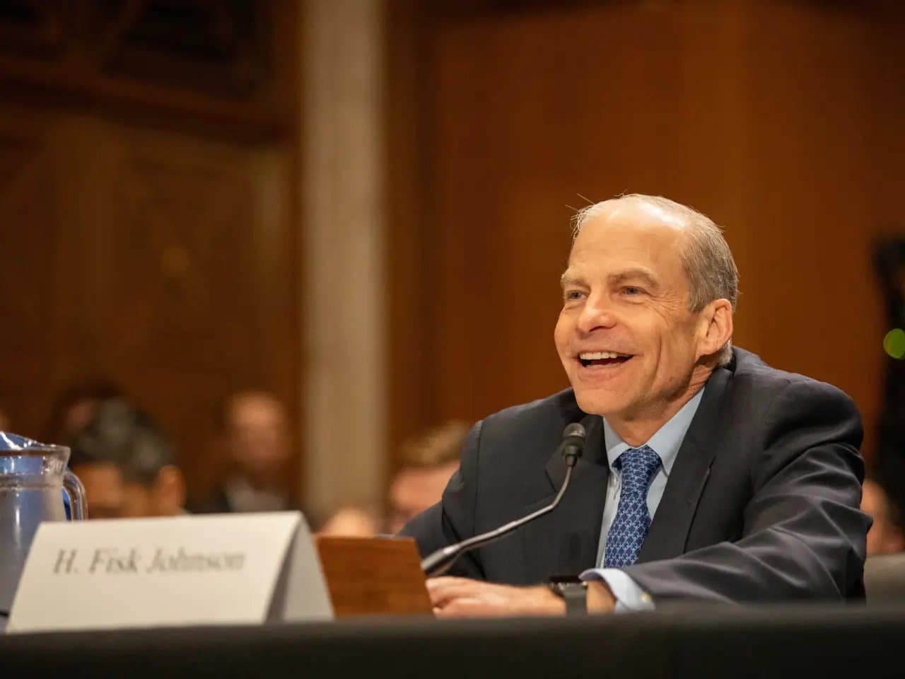 A man in a suit smiles and speaks into a microphone at a conference or hearing. In the foreground, a nameplate reads "H. Fisk Johnson." Others are blurred in the background.