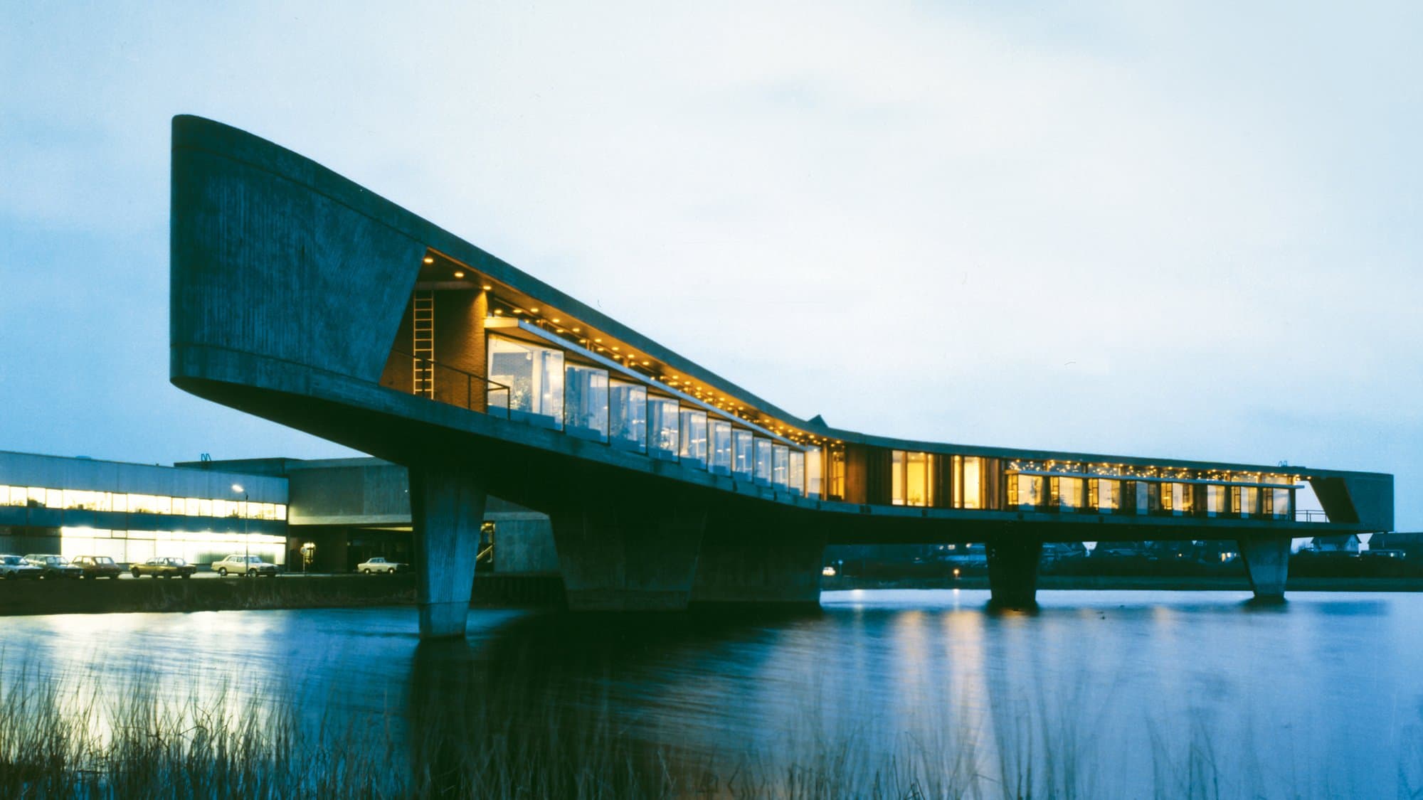 The illuminated concrete structure, designed like a bridge, extends over a body of water at dusk, surrounded by dimly lit buildings and parked cars.