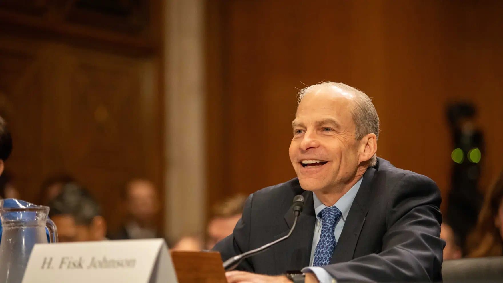 A person sits at a desk speaking into a microphone, smiling. A nameplate reads "H. Fisk Johnson." The setting appears formal, possibly a meeting or hearing, with people blurred in the background.