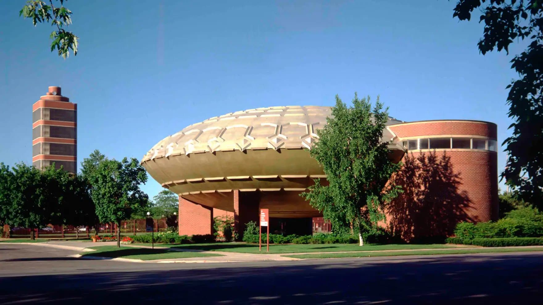 A large, domed building with a unique, UFO-like appearance stands amid trees on a sunny day, with a tall tower visible in the background.