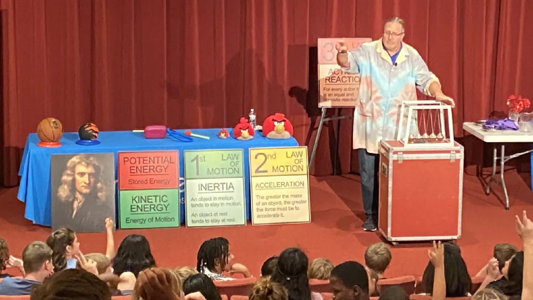 A person conducts a physics demonstration for an audience, with a Newton's cradle and educational posters on motion and energy. Text includes: "POTENTIAL ENERGY Stored Energy," "KINETIC ENERGY Energy of Motion," "1st LAW OF MOTION INERTIA An object in motion tends to stay in motion. An object at rest tends to stay at rest," "2nd LAW OF MOTION ACCELERATION The greater the mass of an object, the greater the force must be to accelerate it," and "3rd LAW ACTION/REACTION For every action there is an equal and opposite reaction." A photo of Isaac Newton is on the table.