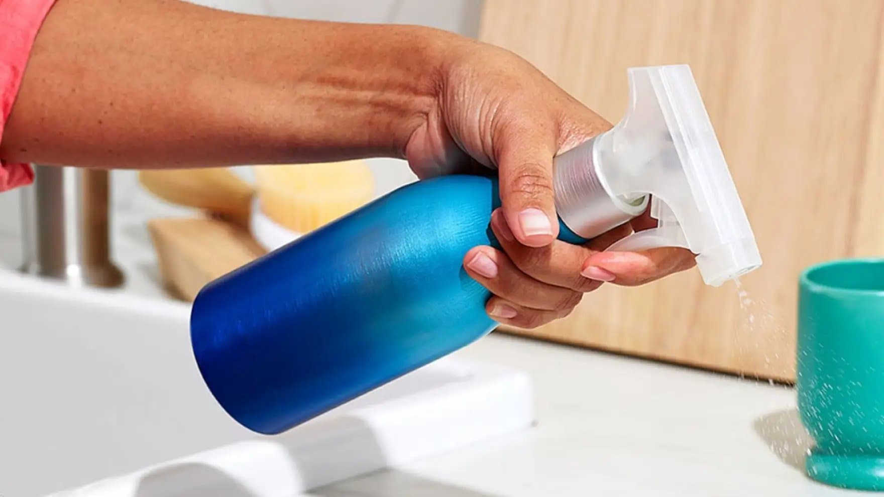 A hand holds a blue spray bottle, squeezing to release liquid, in a kitchen setting with a sink, wooden boards, and a teal cup nearby.