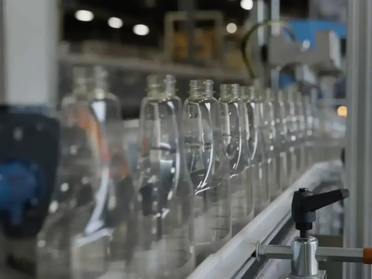 Clear plastic bottles move along a conveyor belt in a factory setting, with machinery and blurred industrial background.