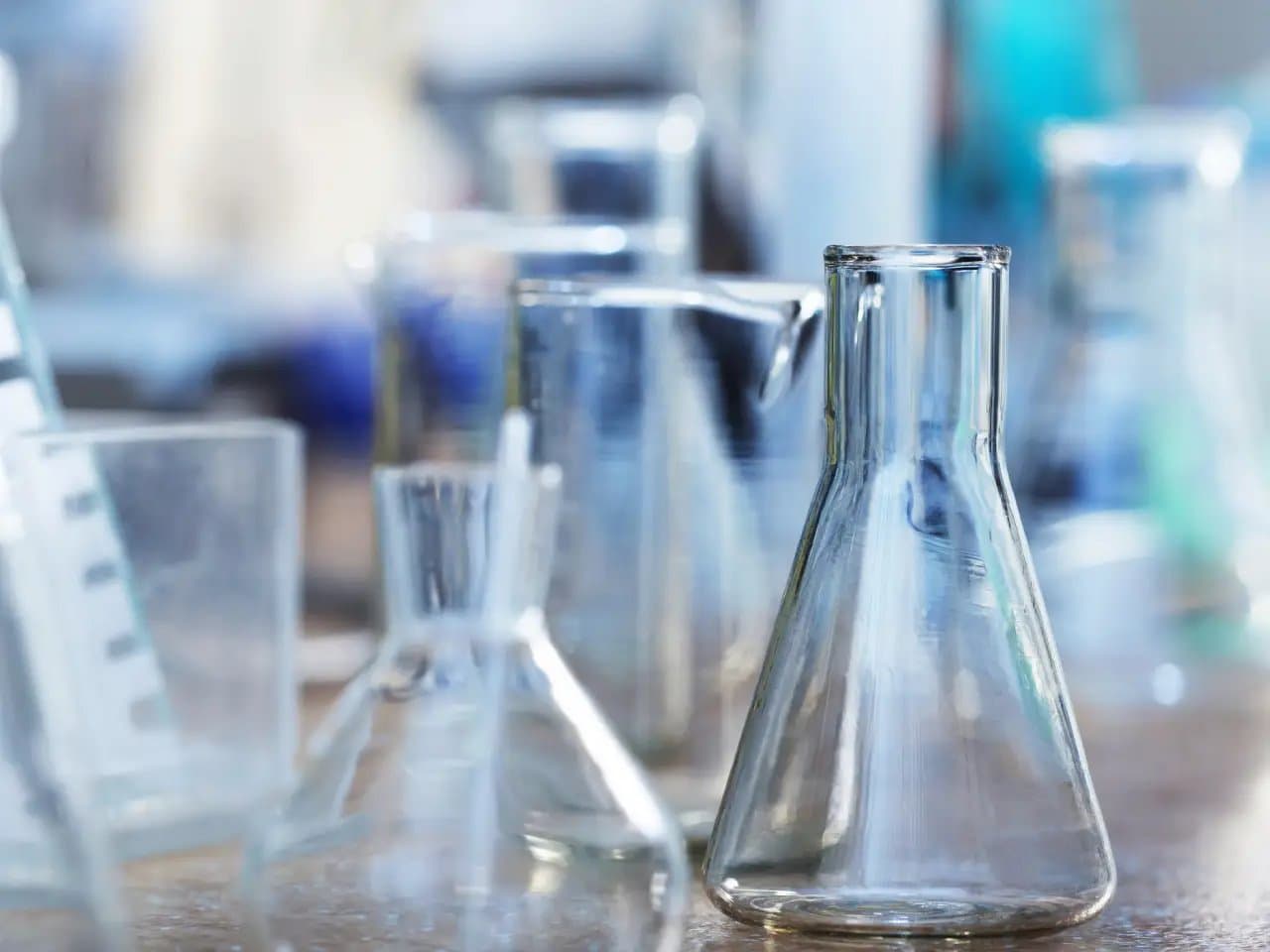 A clear glass flask rests on a laboratory bench among other various glass containers, reflecting light in a scientific workspace.