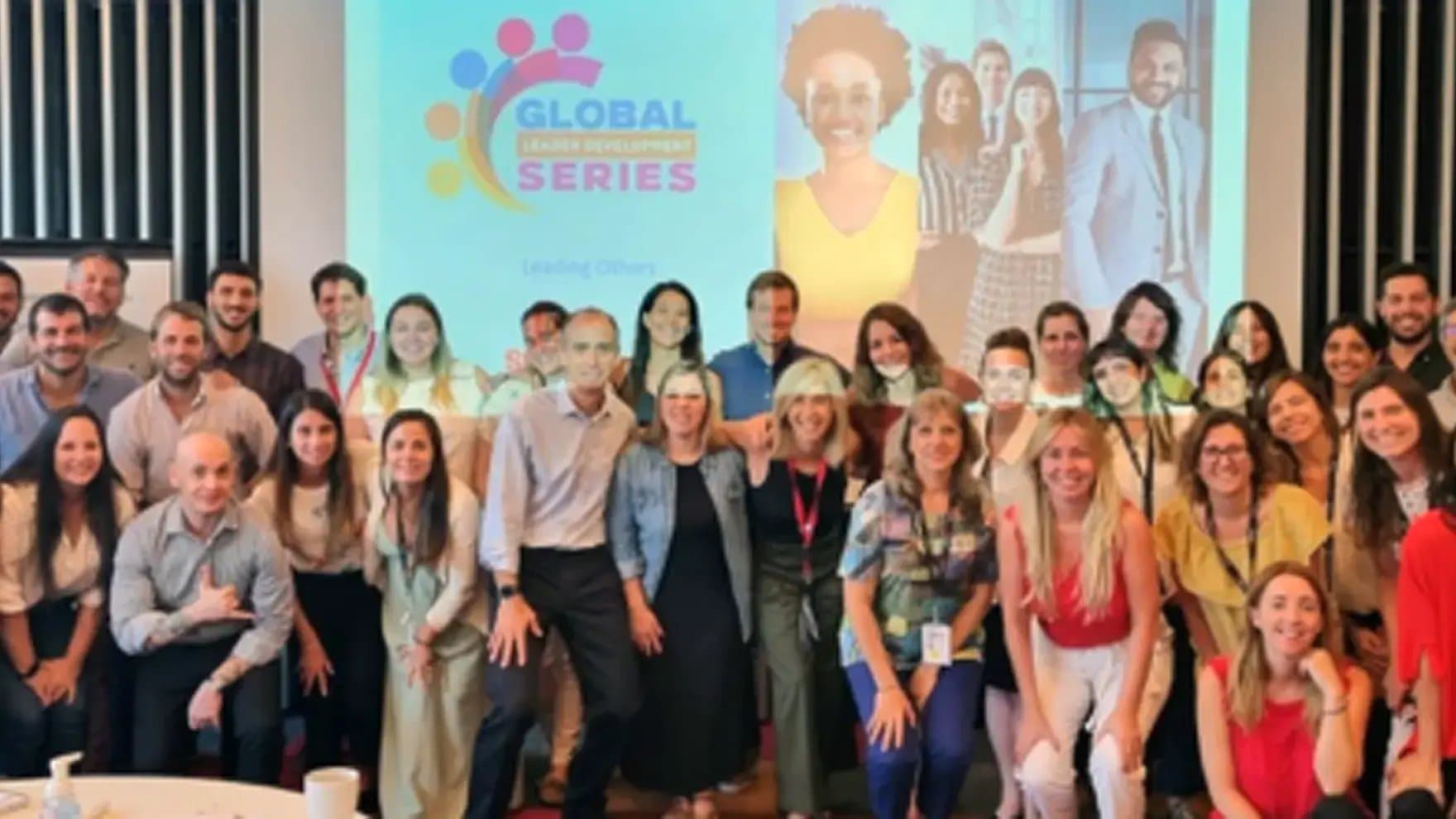 A group of people pose smiling together indoors, in front of a presentation screen displaying "Global Leader Development Series" and diverse individuals' images, suggesting a professional or training event.