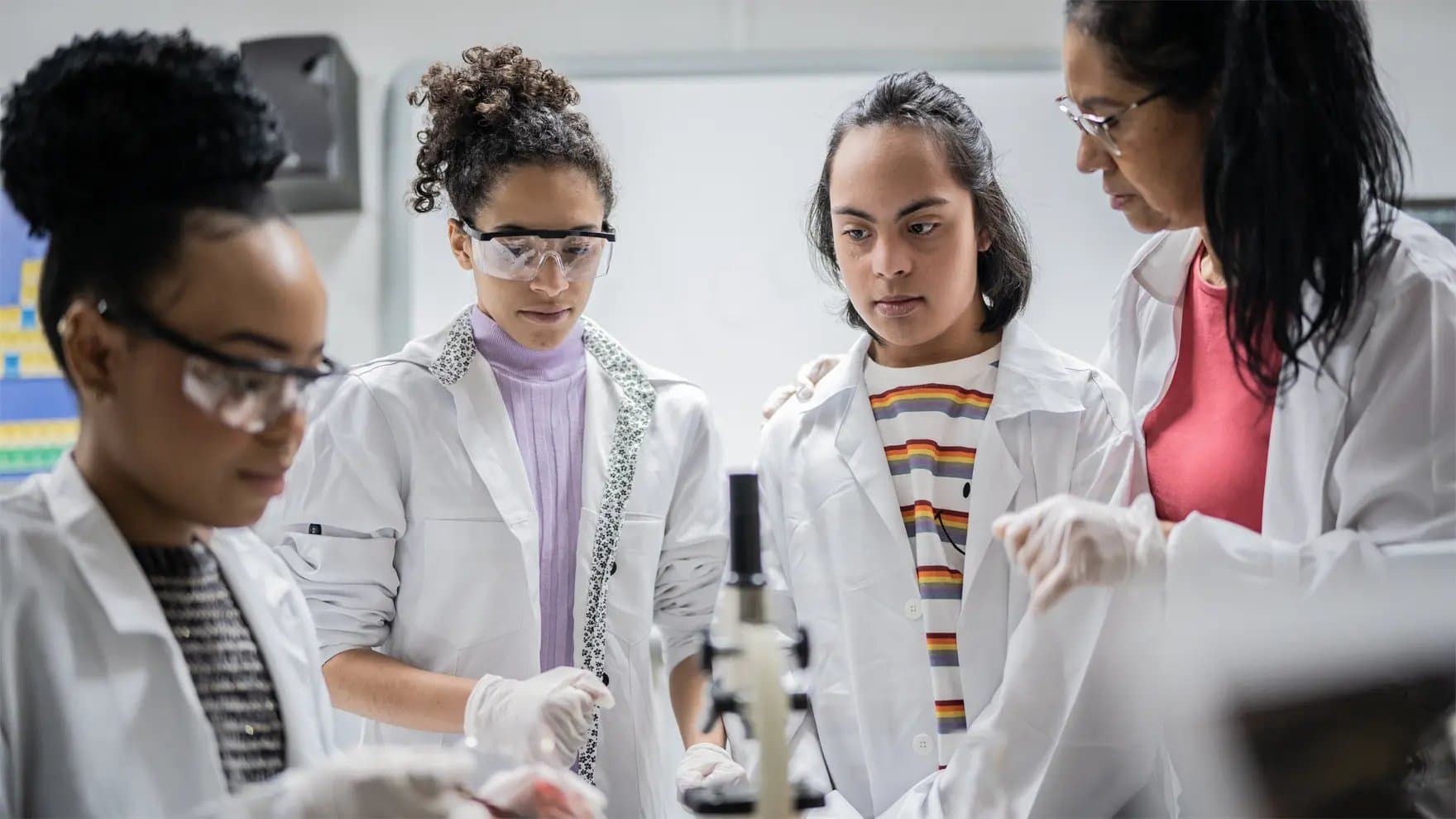 Researchers wearing lab coats and safety goggles observe a microscope in a laboratory setting, focusing intently on their work, surrounded by scientific equipment in a classroom-like environment.