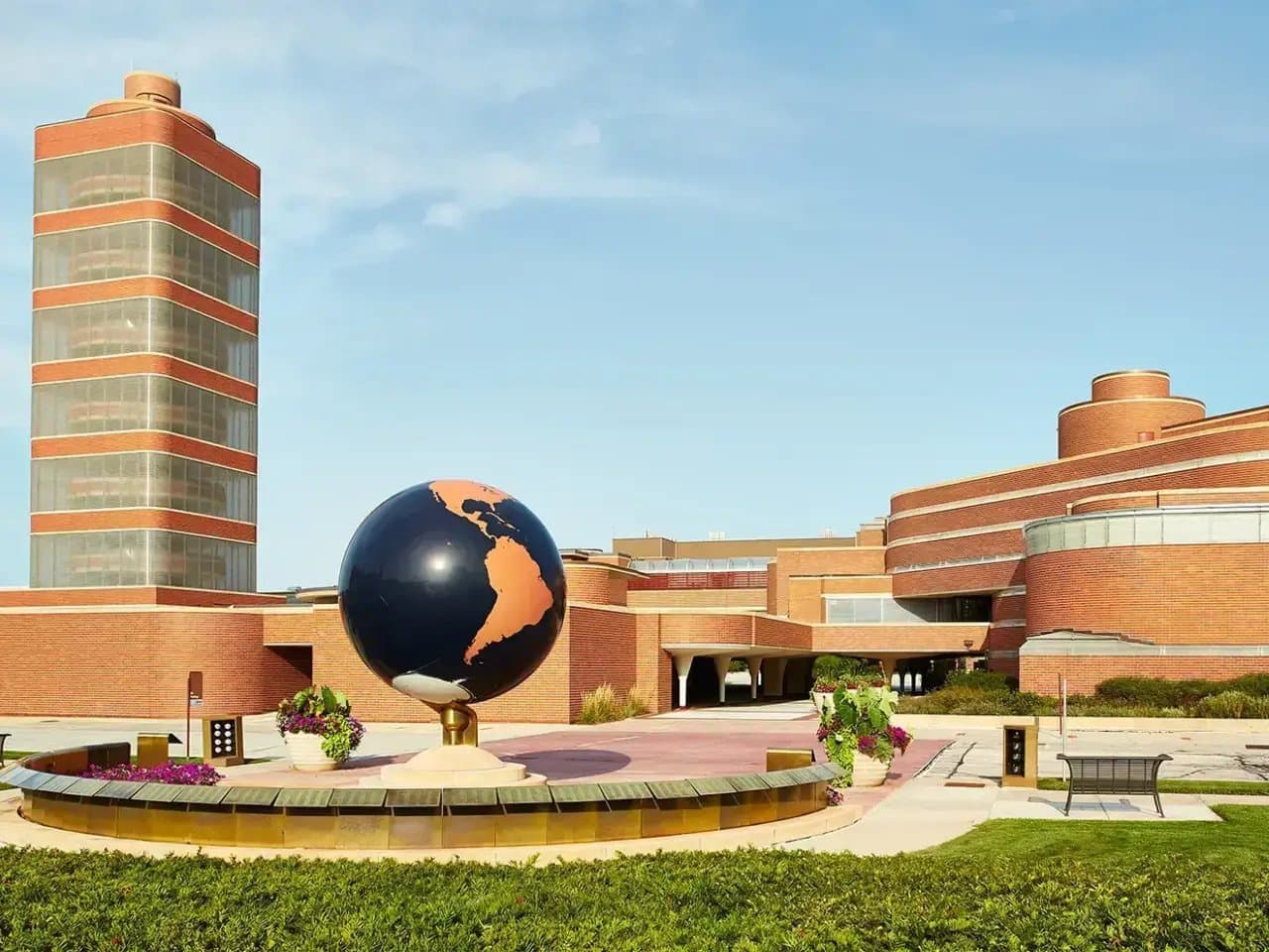 A large globe sculpture sits on a circular fountain, surrounded by colorful plants and benches, outside a modern brick building with a tall cylindrical tower.