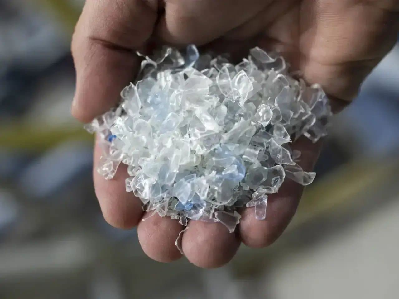 A hand holds translucent plastic flakes, centered in the frame. The context is a blurred, possibly industrial or recycled material background, suggesting a focus on recycling or processing.