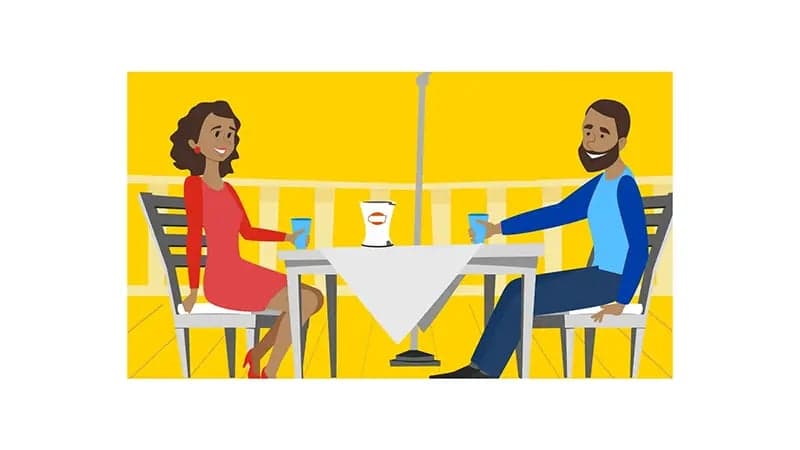 A man and a woman sit across from each other at a table, holding drinks, with a yellow background and a small container labeled "Lipton" on the table.