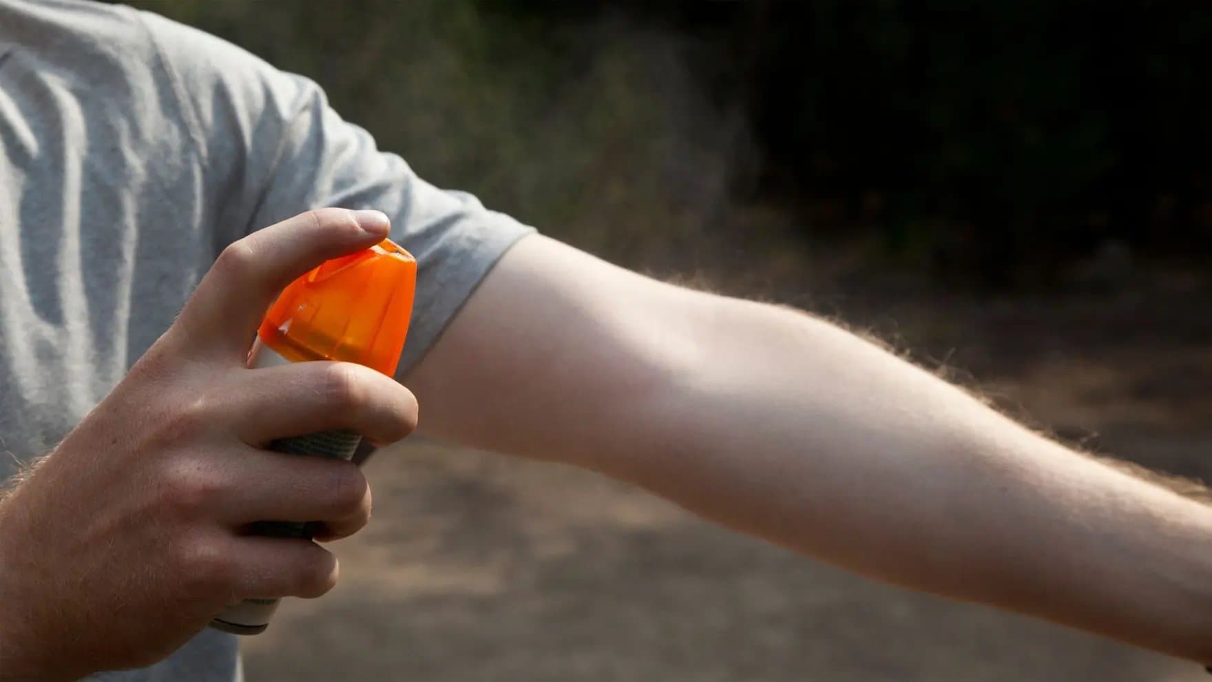 A person sprays an arm with insect repellent from an orange-topped can, outdoors in a wooded area.