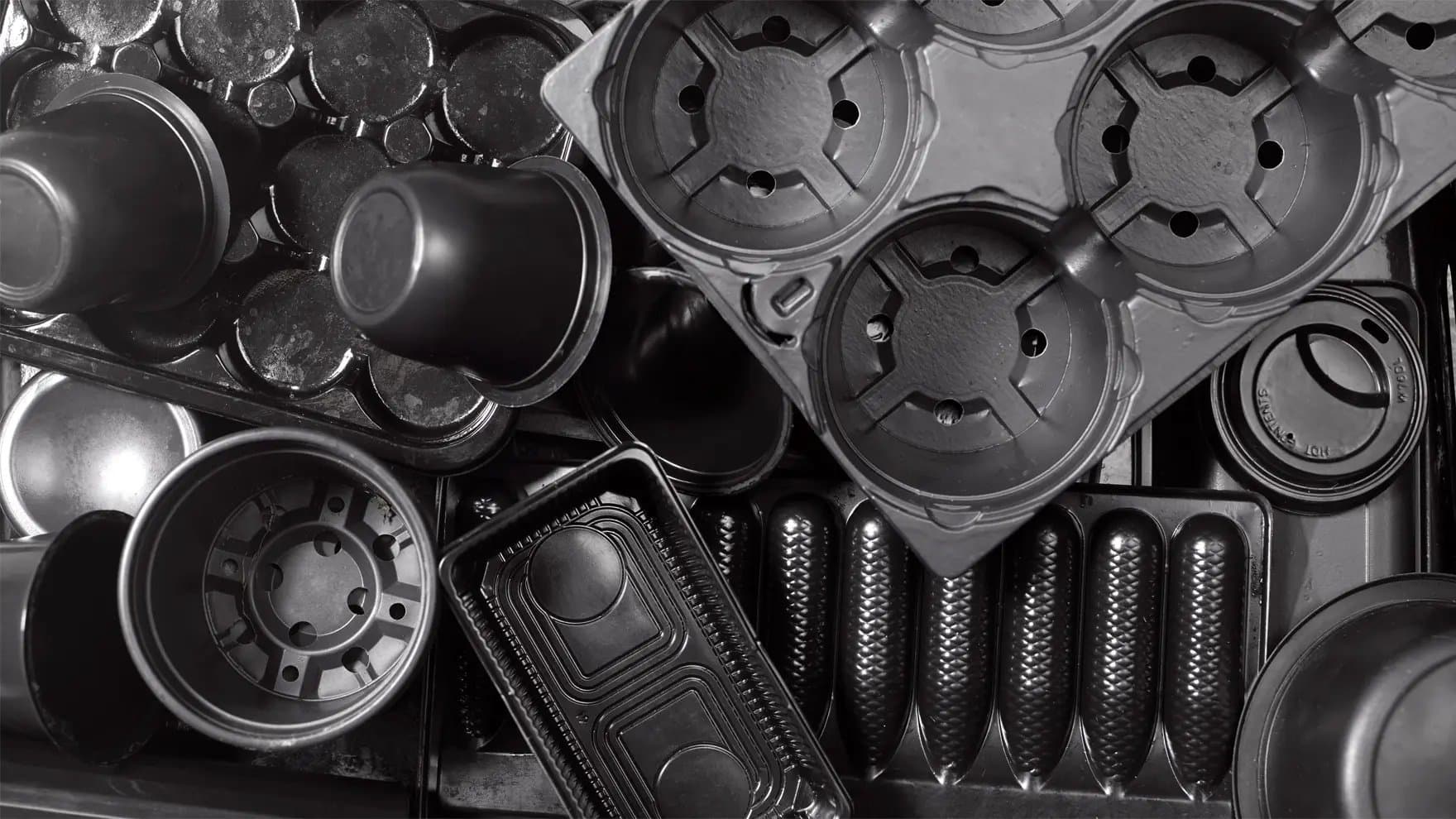 A collection of black plastic containers and lids, including cups, trays, and packaging materials, overlap each other in a close, cluttered arrangement.