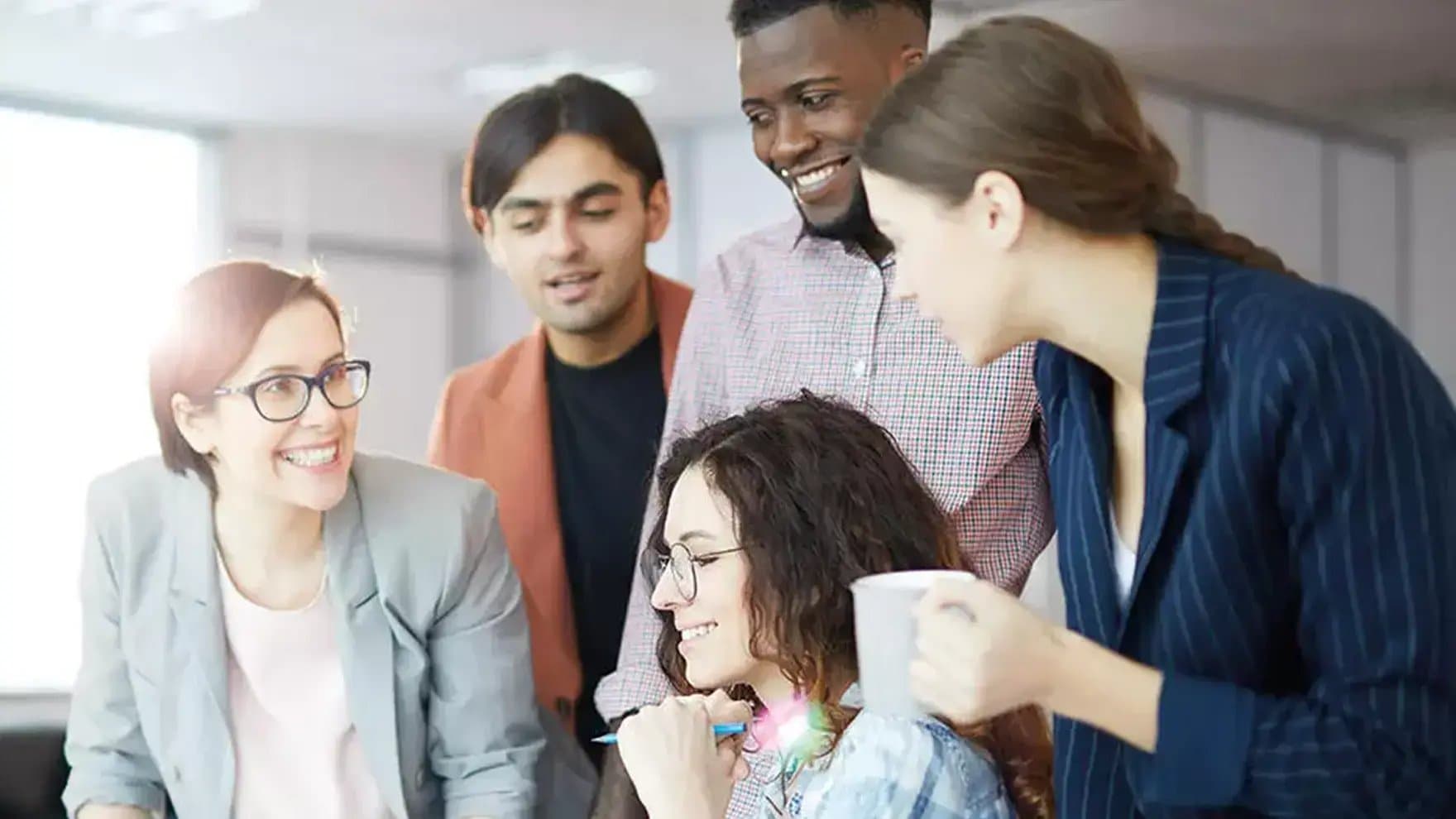 A group of five people are smiling and discussing something, gathered around a seated woman holding a pen. They appear to be in a modern office setting.