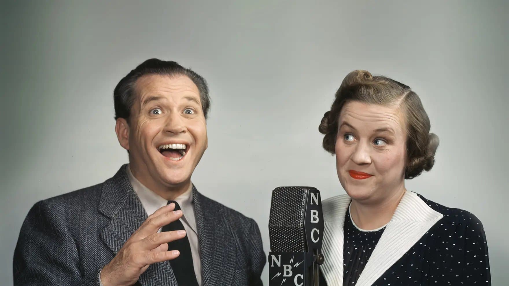 Two people stand excitedly near an NBC microphone. The man is gesturing with a wide smile, while the woman glances sideways with a smirk. The background is plain gray.