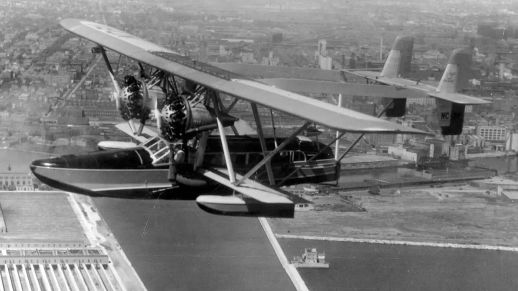 A biplane seaplane flying over a cityscape, featuring twin engines and a distinctive cockpit, with urban buildings and a waterfront below, indicating an early aviation era.