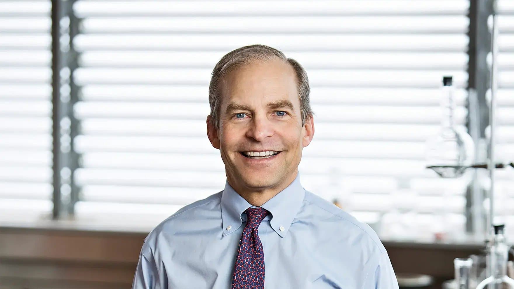 A person is smiling while wearing a light blue shirt and patterned tie. They stand in front of a window with horizontal blinds, with laboratory equipment visible in the background.