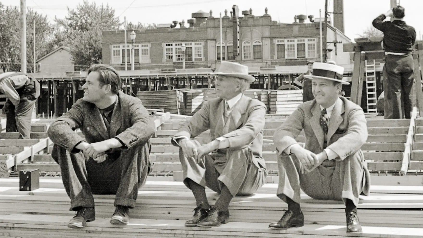 Three men sit on wooden planks at a construction site, wearing suits and hats. In the background, buildings and workers add to the busy urban scene.