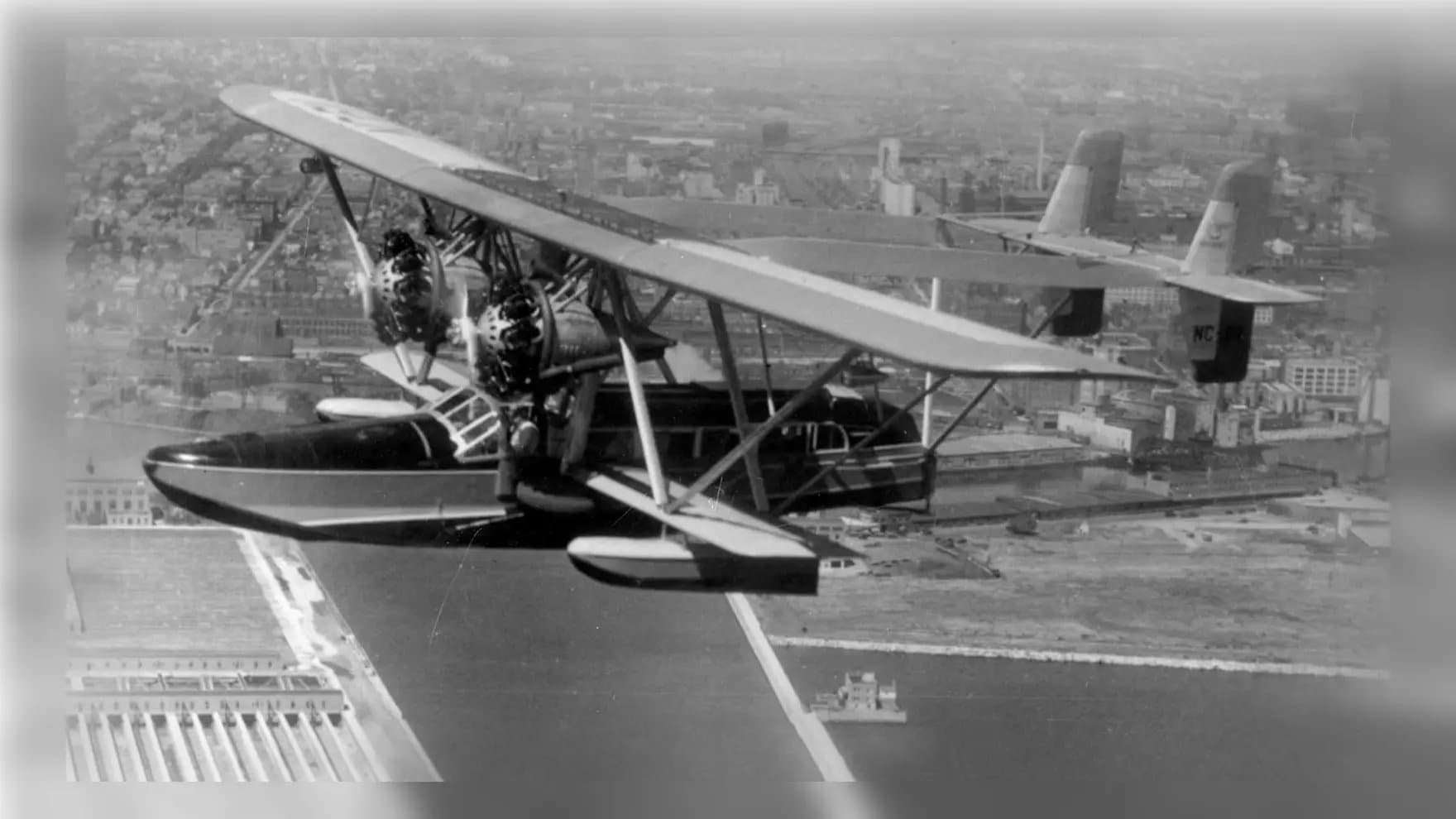 A vintage biplane flies over a cityscape, showcasing its twin engines and pontoons. Urban buildings and waterways create a detailed backdrop beneath the aircraft.