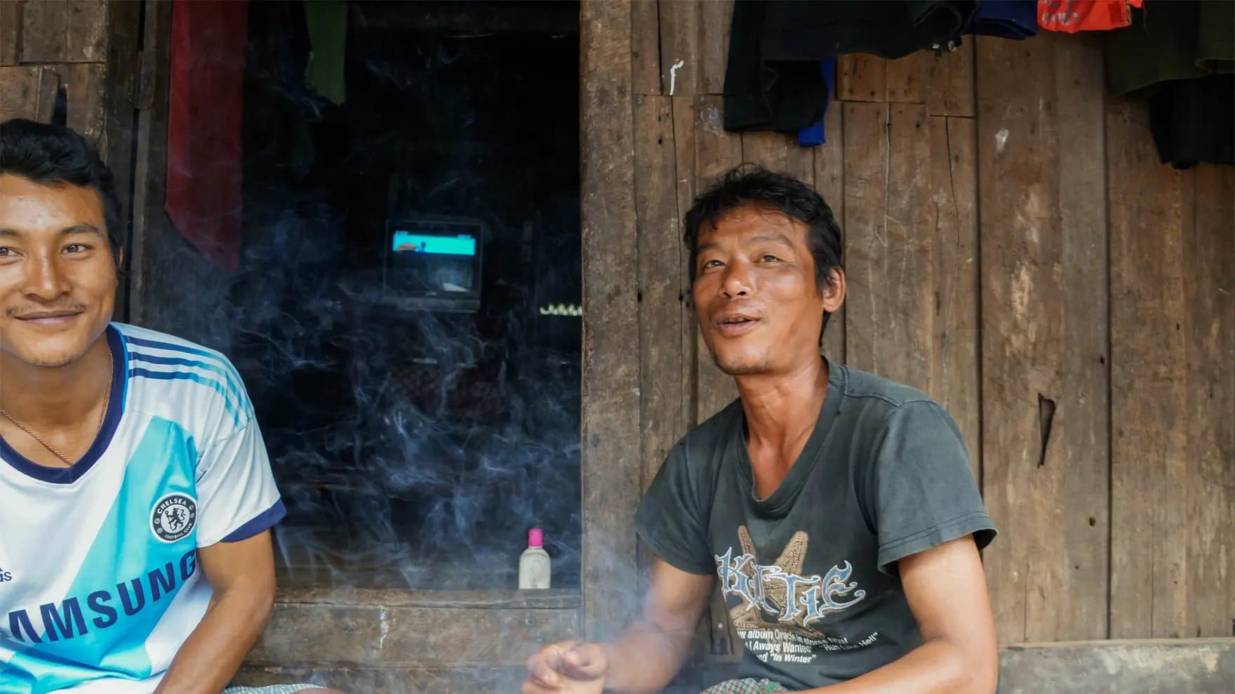 Two men sit smiling in front of a wooden wall. Smoke swirls around them, and a small TV is visible inside the dimly lit room. Clothing hangs above.