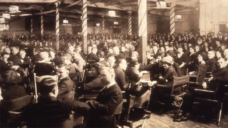 A large group of people sit and converse in a crowded hall with wooden chairs, surrounded by decorative columns and overhead lights, creating a formal gathering atmosphere.