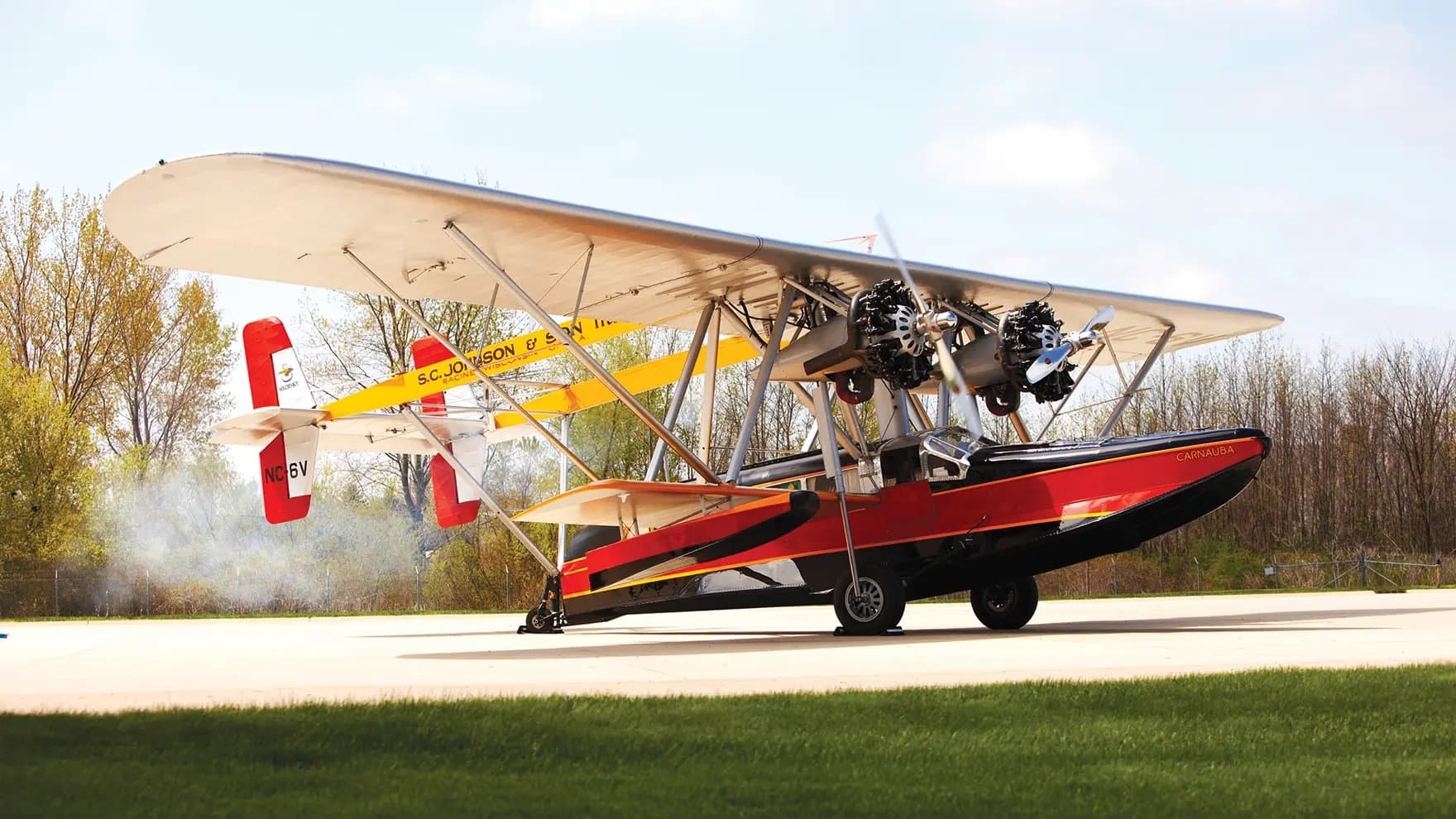 A vintage seaplane, named "Carnauba," idles on a sunny tarmac with smoke trailing. Surrounding trees create a green backdrop under a clear sky. The tail reads "S.C. Johnson & Son, Inc."