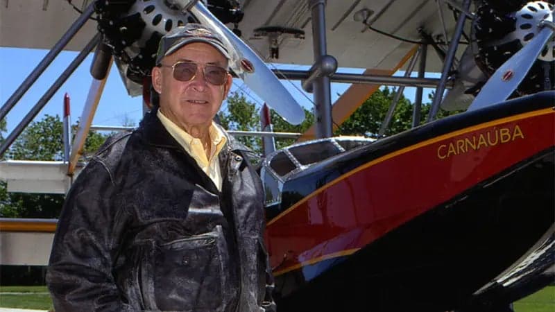 A person wearing a leather jacket and cap stands beside a vintage airplane with visible propellers. The aircraft is red and black with "CARNAUBA" written on the side, set outdoors.