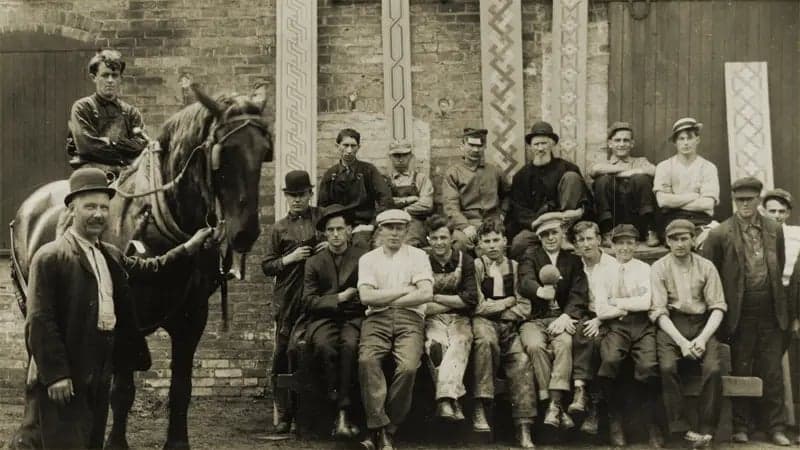 A group of workers pose in front of a brick building. One man sits on a horse while others stand or lean casually, some wearing hats and work clothes.