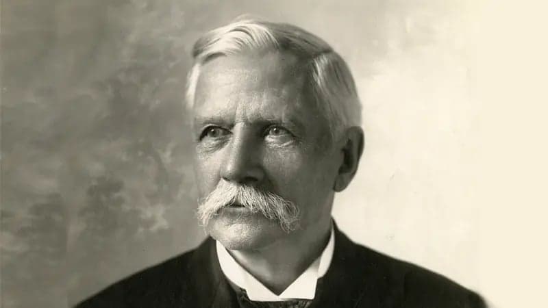 Elderly man with a thick mustache gazes upward, wearing a formal suit, in a sepia-toned portrait against a textured background.