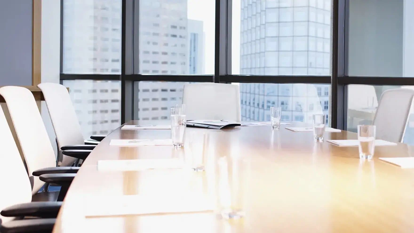 A conference table holds glass cups and documents in a modern office with large windows. Skyscrapers are visible through the windows, enhancing the professional setting.