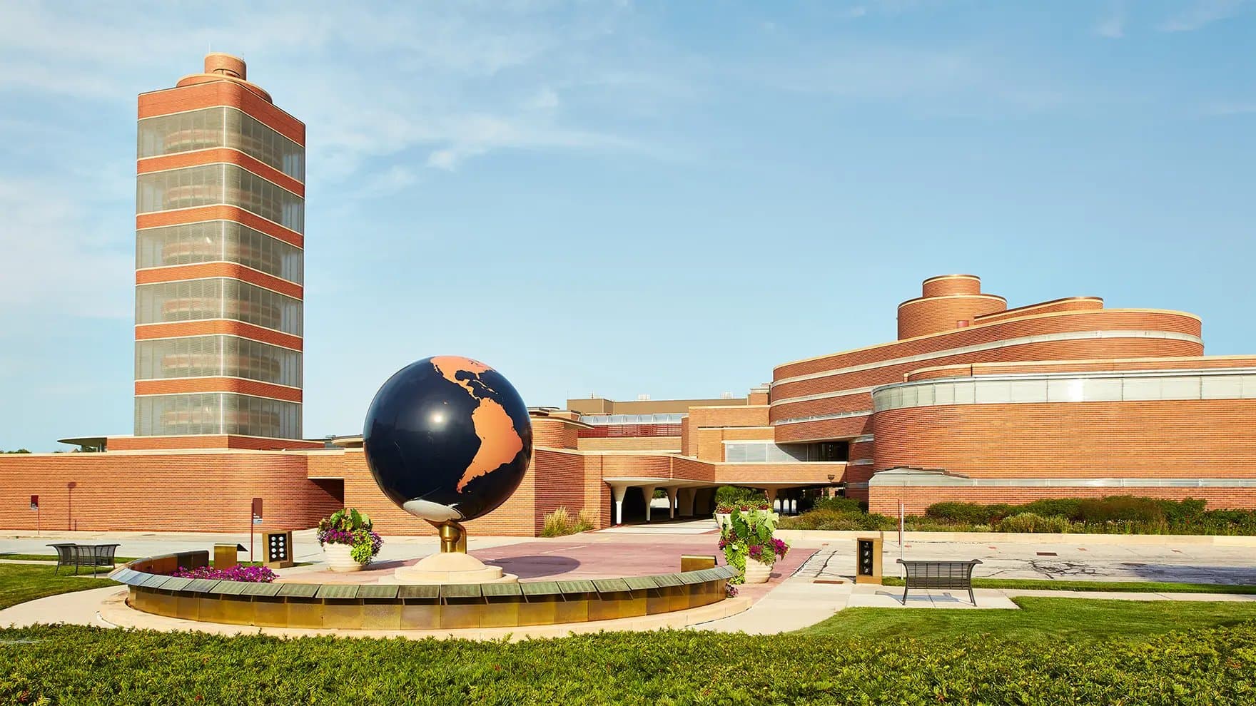 A large globe sculpture stands on a pedestal, surrounded by landscaped gardens, in front of a modern, brick building featuring a tall, cylindrical tower under a clear blue sky.