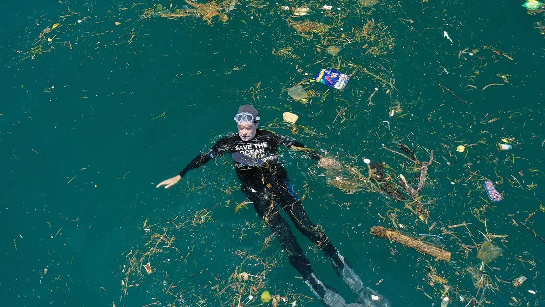 A swimmer, wearing a shirt that says "SAVE THE OCEAN," floats among debris and litter in a body of water.