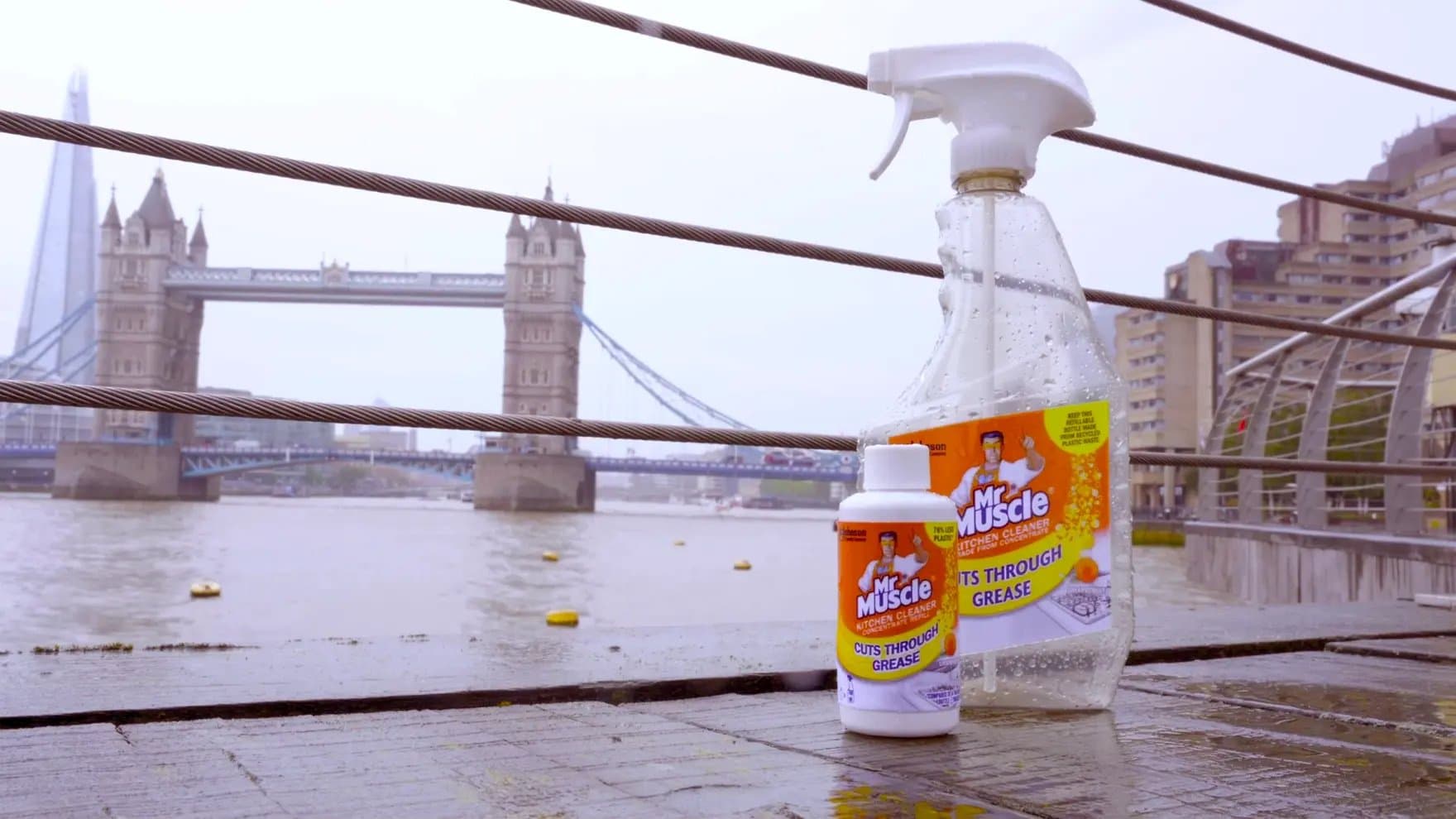 A bottle of "Mr Muscle Kitchen Cleaner" stands beside a spray bottle on a wet boardwalk, overlooking Tower Bridge and the river, with buildings in the background.