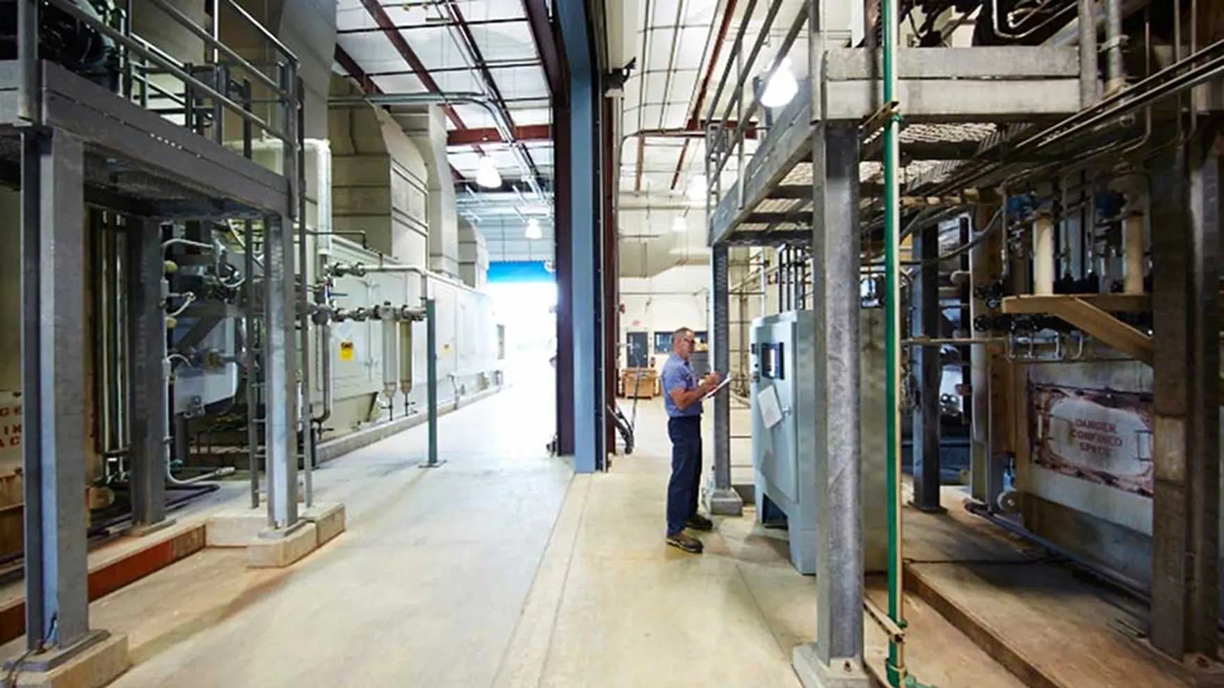 A worker inspects machinery, taking notes inside an industrial facility. Piping and control panels surround the area, with "DANGER CORROSIVE SPRAY" marked on a visible sign.