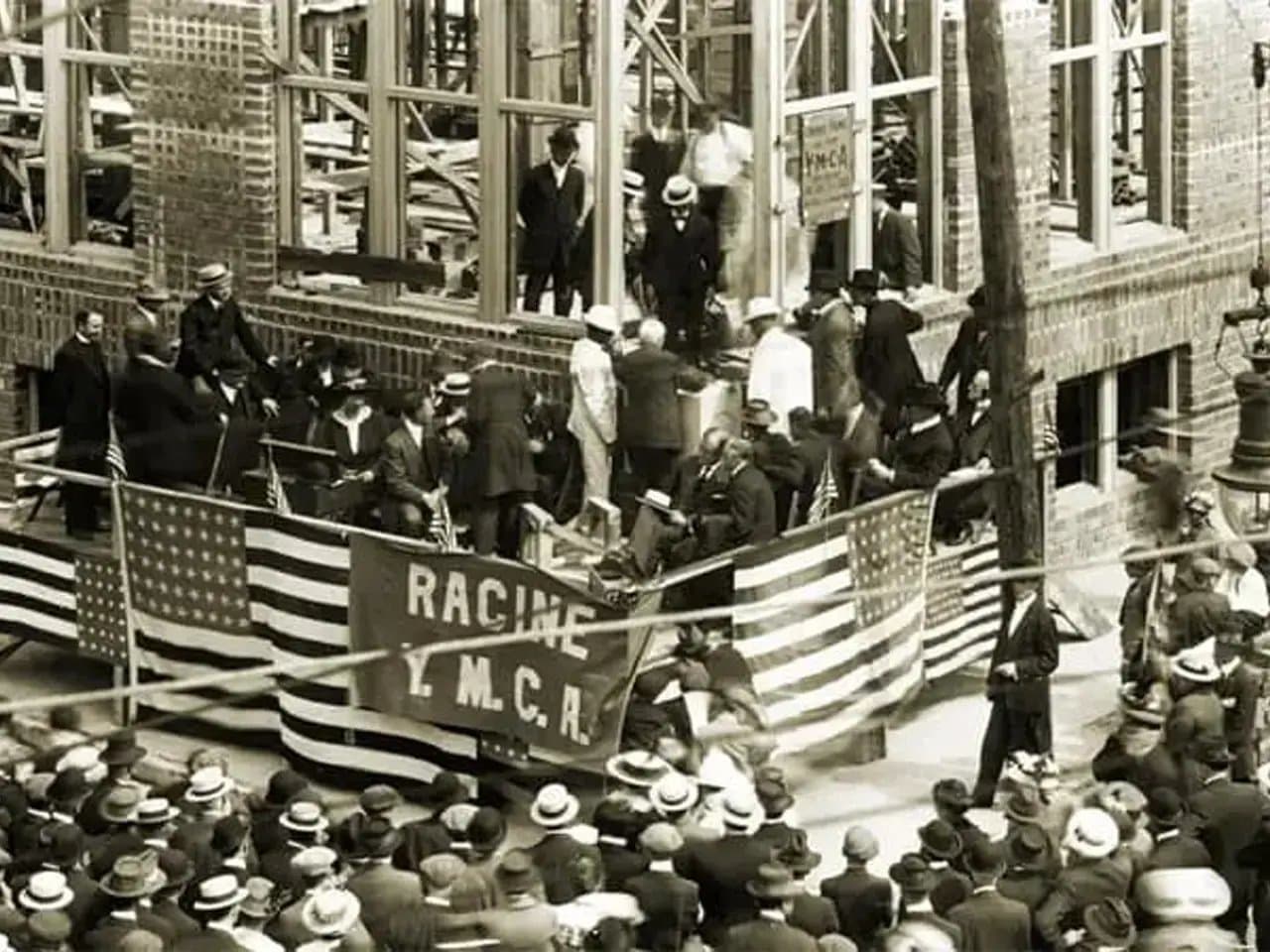 Exterior view of brick building with crowd of men in suits and hats with American flags and sign that reads "Racine Y.M.C.A"