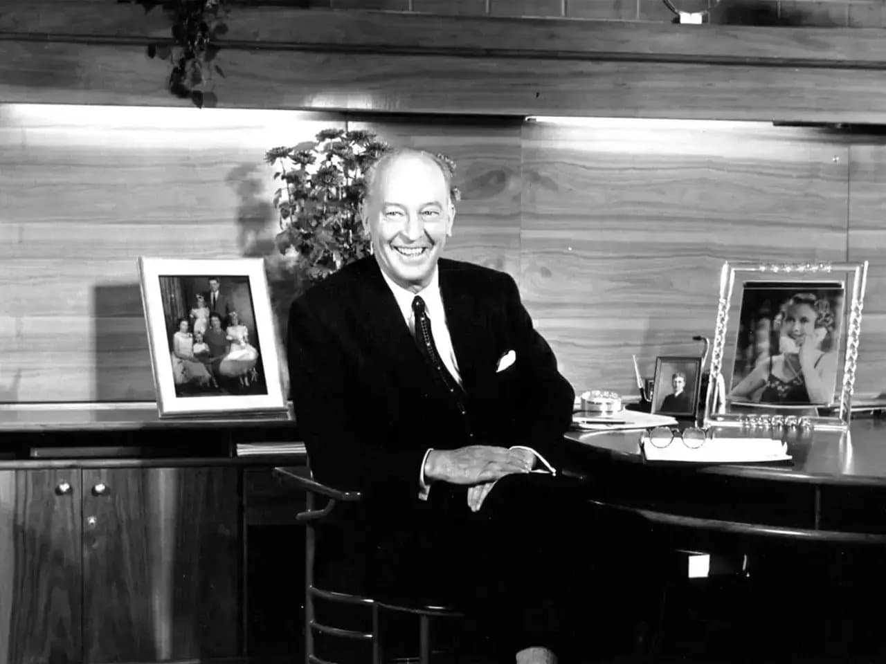 A man in a suit sits smiling at a desk, adorned with family photographs and a plant, set against a wood-paneled background.