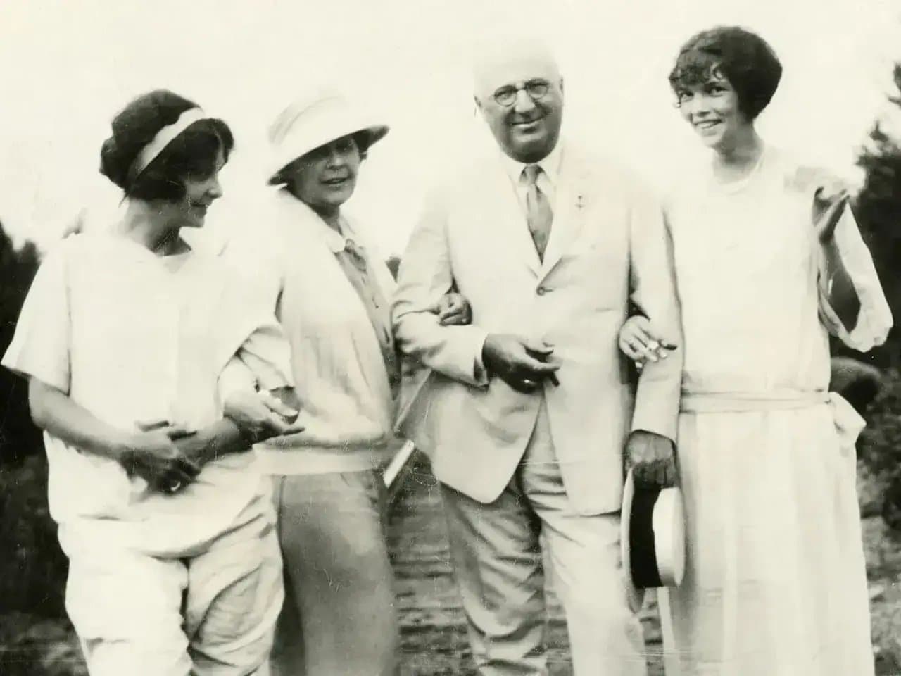 Four people stand together, smiling warmly. The central figure, a man in a suit holding a hat, is flanked by women in casual attire, suggesting a leisurely outdoor setting.
