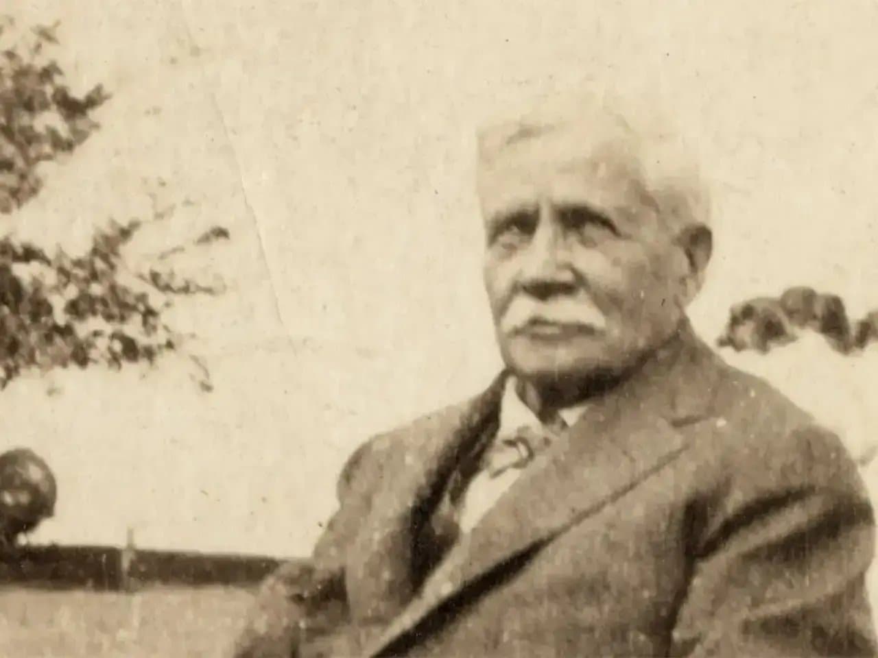 An elderly man with a mustache sits outdoors, wearing a suit, with trees and open skies in the background, creating an early 20th-century atmosphere.