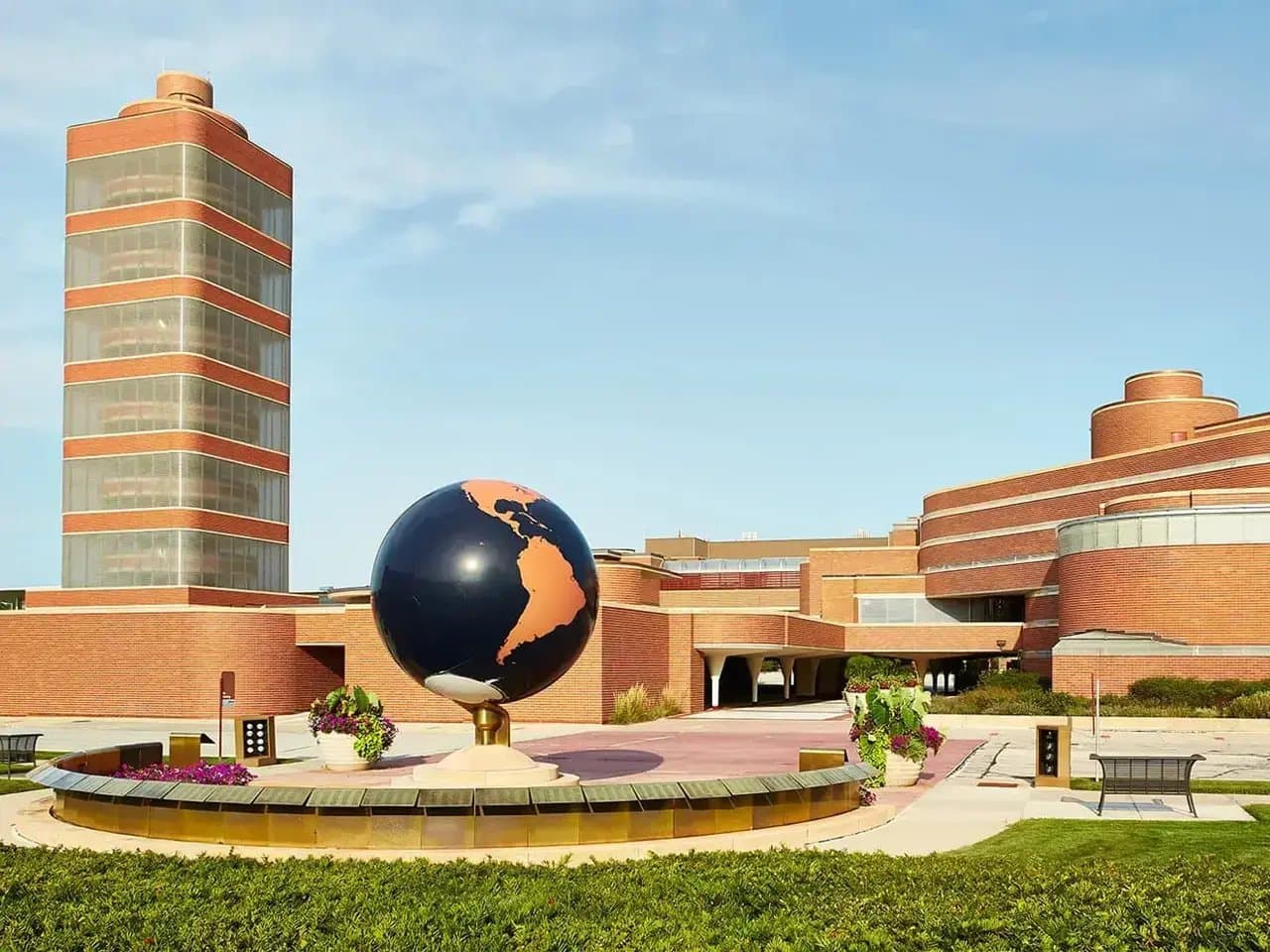 A large globe sculpture sits on a pedestal in a plaza surrounded by brick buildings. Potted plants and benches enhance the open space under a clear blue sky.