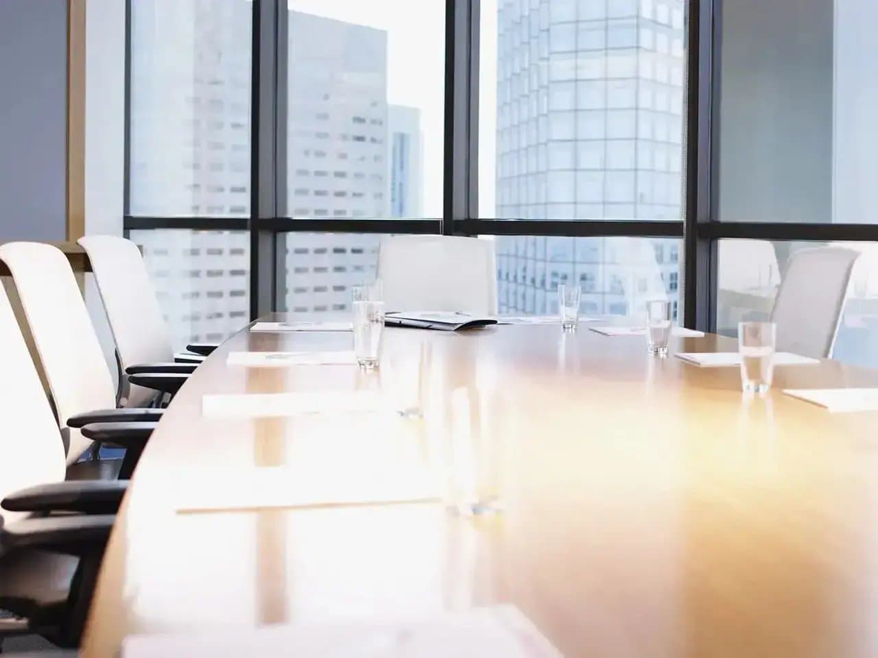 A large wooden conference table, surrounded by white chairs, holds scattered papers and glasses of water, situated inside a bright room with floor-to-ceiling windows overlooking city skyscrapers.