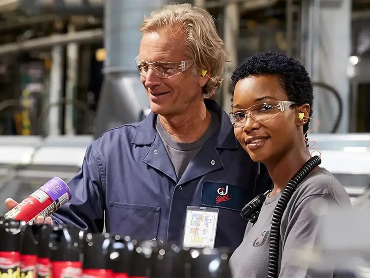 Two workers, wearing safety glasses and ear protection, inspect a canister in a factory setting, surrounded by various bottles on a production line.