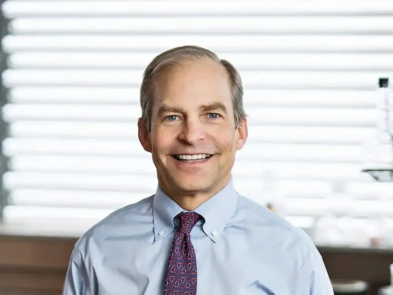 A man in a light blue shirt and patterned tie smiles confidently, standing indoors with blurred horizontal blinds in the background.