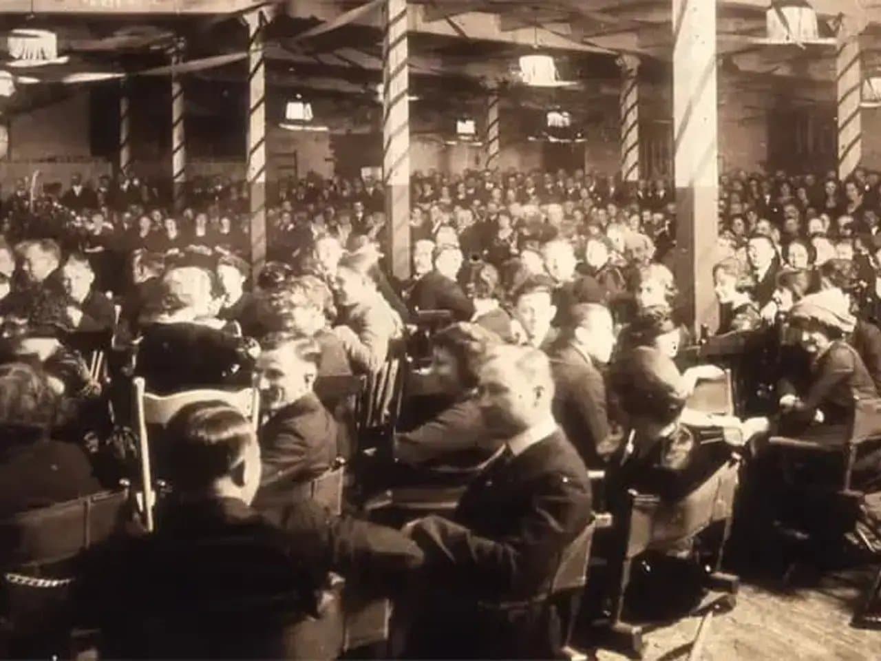 A large crowd is seated in a dimly lit, indoor hall filled with rows of chairs, facing forward, suggesting an event or gathering. The atmosphere is busy and expectant.