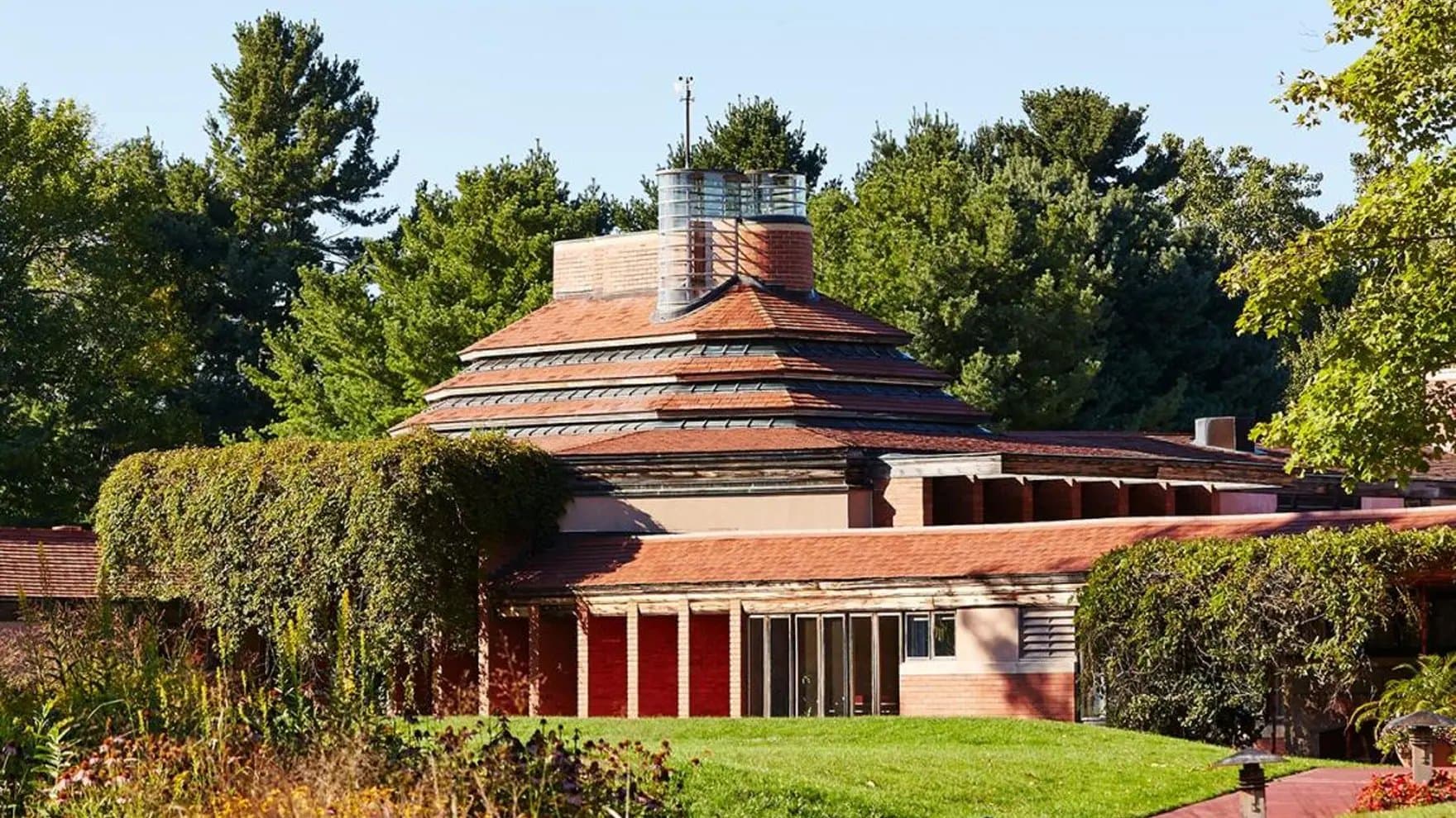 A multi-tiered building with a terracotta roof and glass chimney stands amidst lush greenery, surrounded by trees and flowering plants under a clear blue sky.