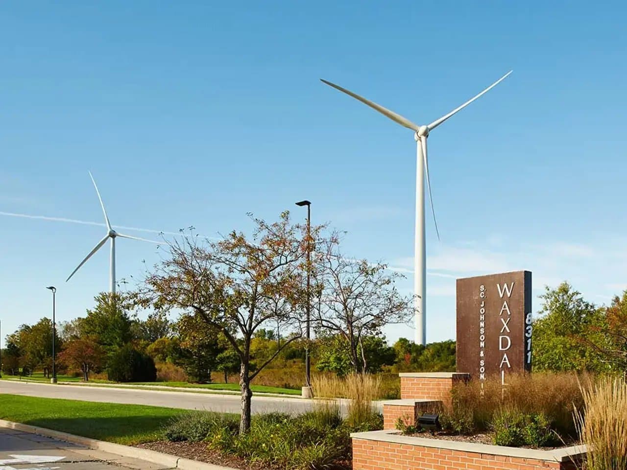 Two wind turbines stand erect on a clear day near a road lined with trees. A sign reads, "S.C. Johnson & Son, Inc. Waxdale 8311."
