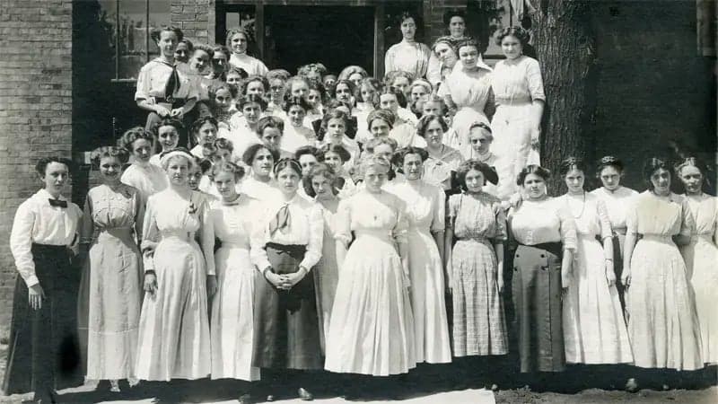 A large group of women, dressed in early 20th-century attire, stands closely together for a group photo. They are outdoors, near a brick building and a large tree.