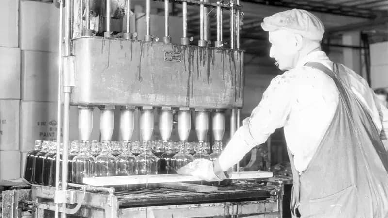 A worker operates a bottling machine, filling glass bottles with liquid in an industrial setting. Cardboard boxes stack in the background of the factory environment.